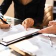 Close-up Of Two Businesspeople Hand Working On Contract Paper Over Desk