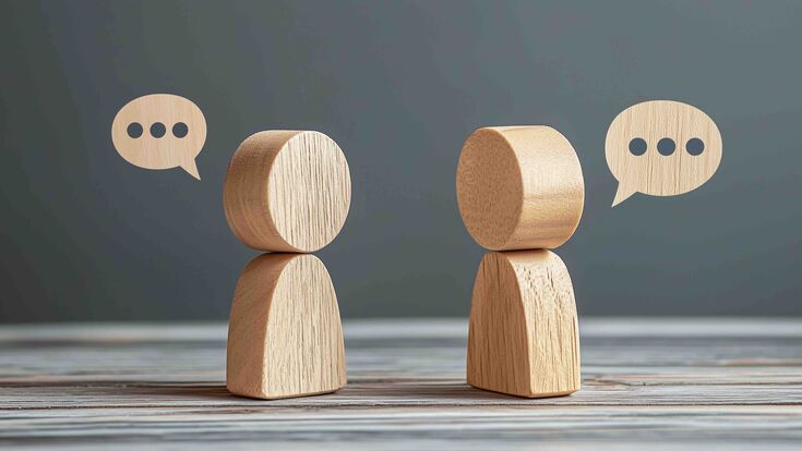 Two wooden figures engaged in a conversation with speech bubbles on a dark gray background, symbolizing dialogue and interaction with a minimalist aesthetic.