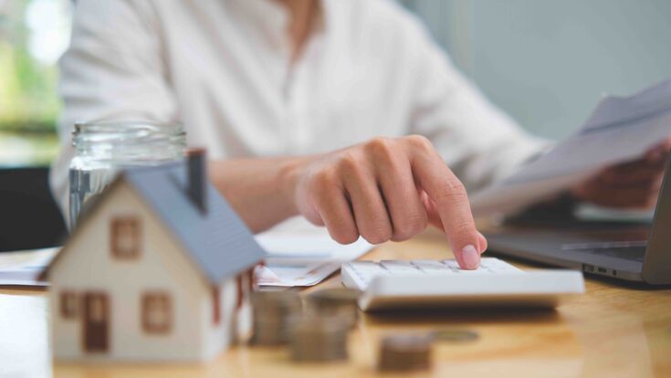Real estate investment and mortgage calculation concept with businessman using calculator, coins stacked on desk, laptop, and small house model, representing property finance, budget planning, and home