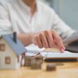 Real estate investment and mortgage calculation concept with businessman using calculator, coins stacked on desk, laptop, and small house model, representing property finance, budget planning, and home